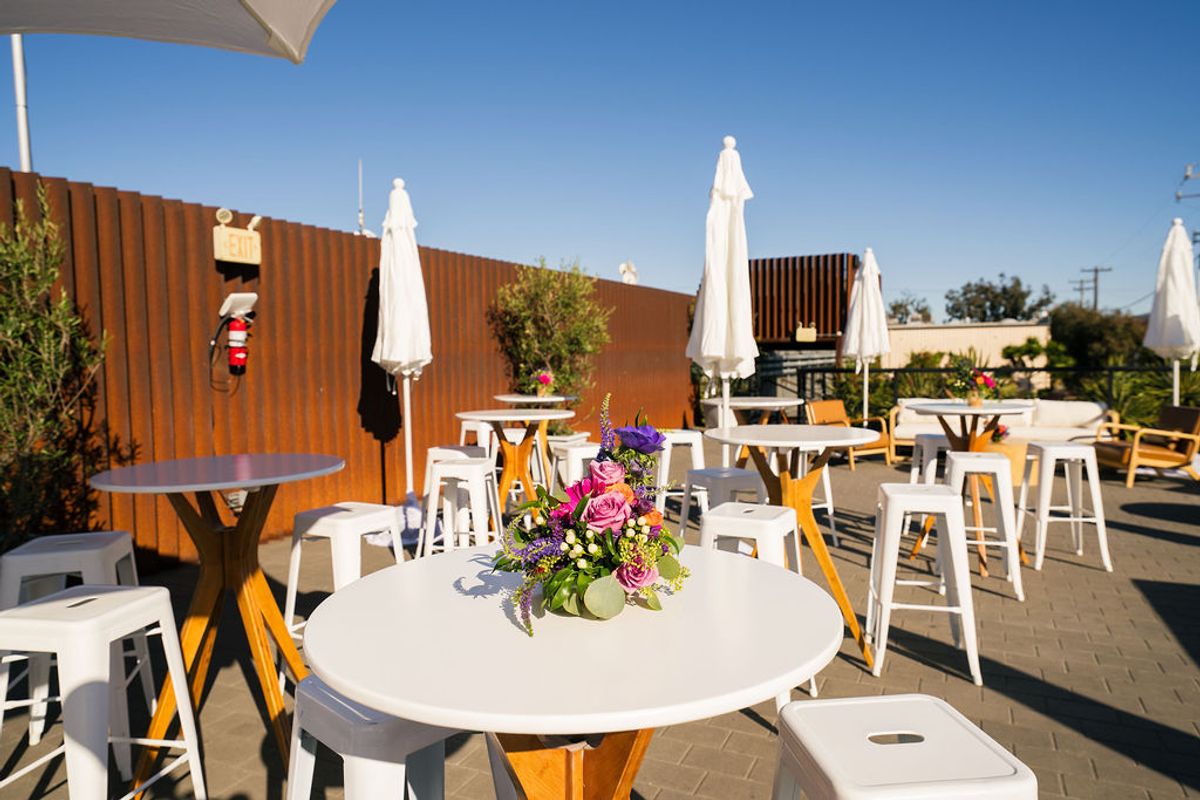 Outdoor patio with white tables, stools, closed umbrellas, and purple flower centerpieces under clear blue sky
