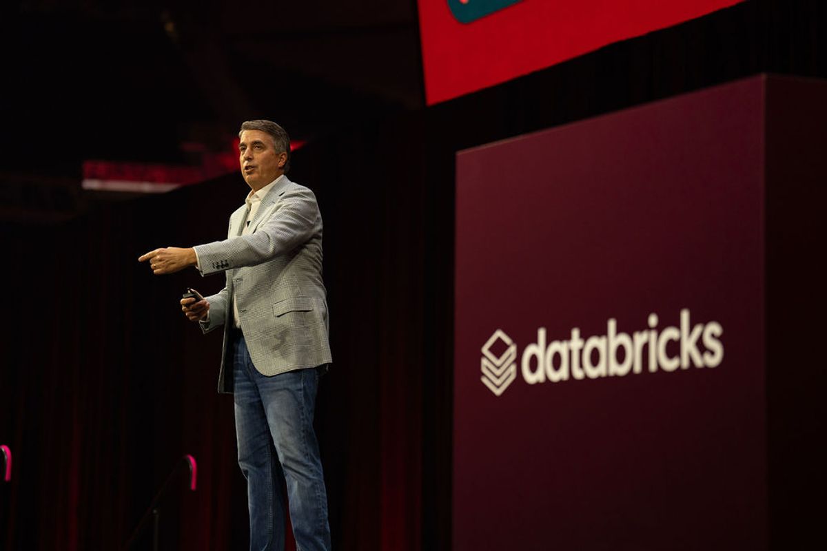 Speaker in gray blazer presenting on stage with Databricks logo backdrop at conference venue