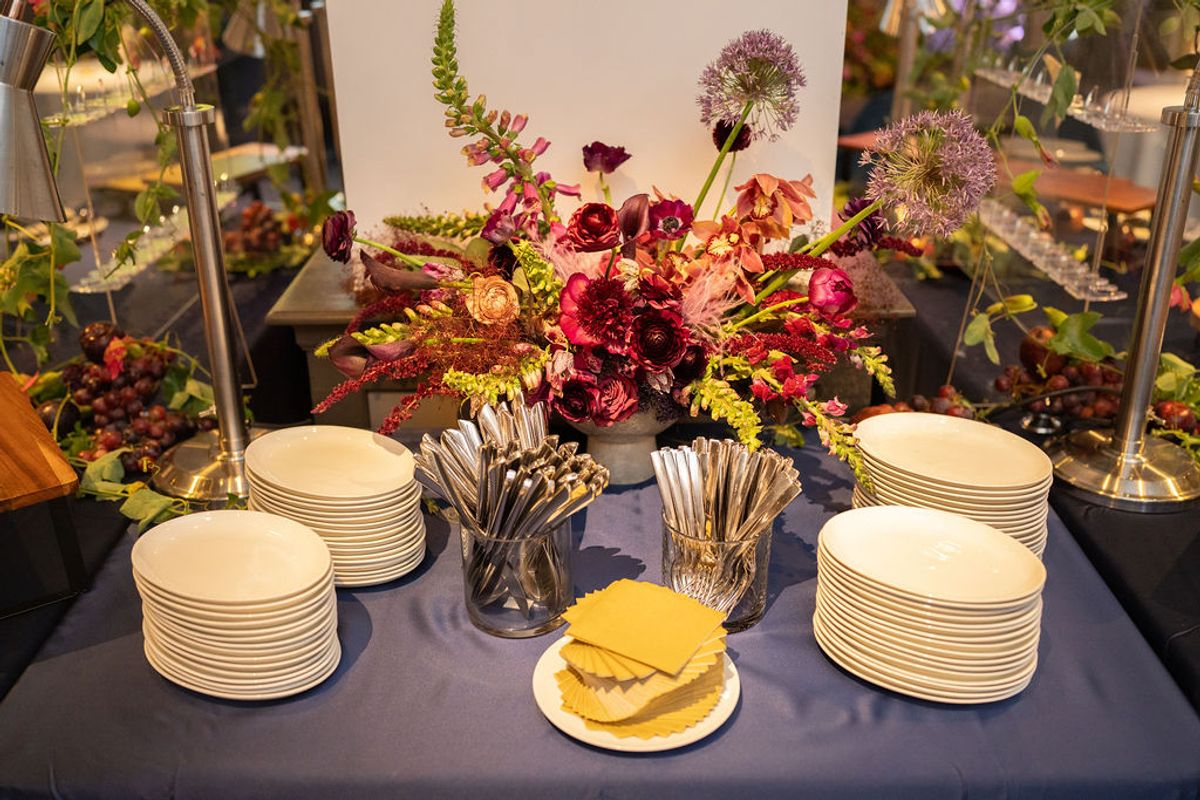 Buffet table with stacked plates, utensils, floral centerpiece, and food warmers at networking reception