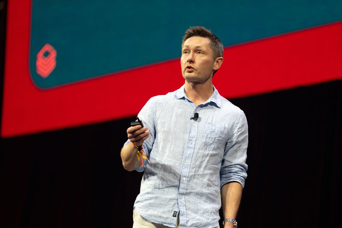 Speaker in light blue shirt holding microphone presents on stage with red and teal backdrop