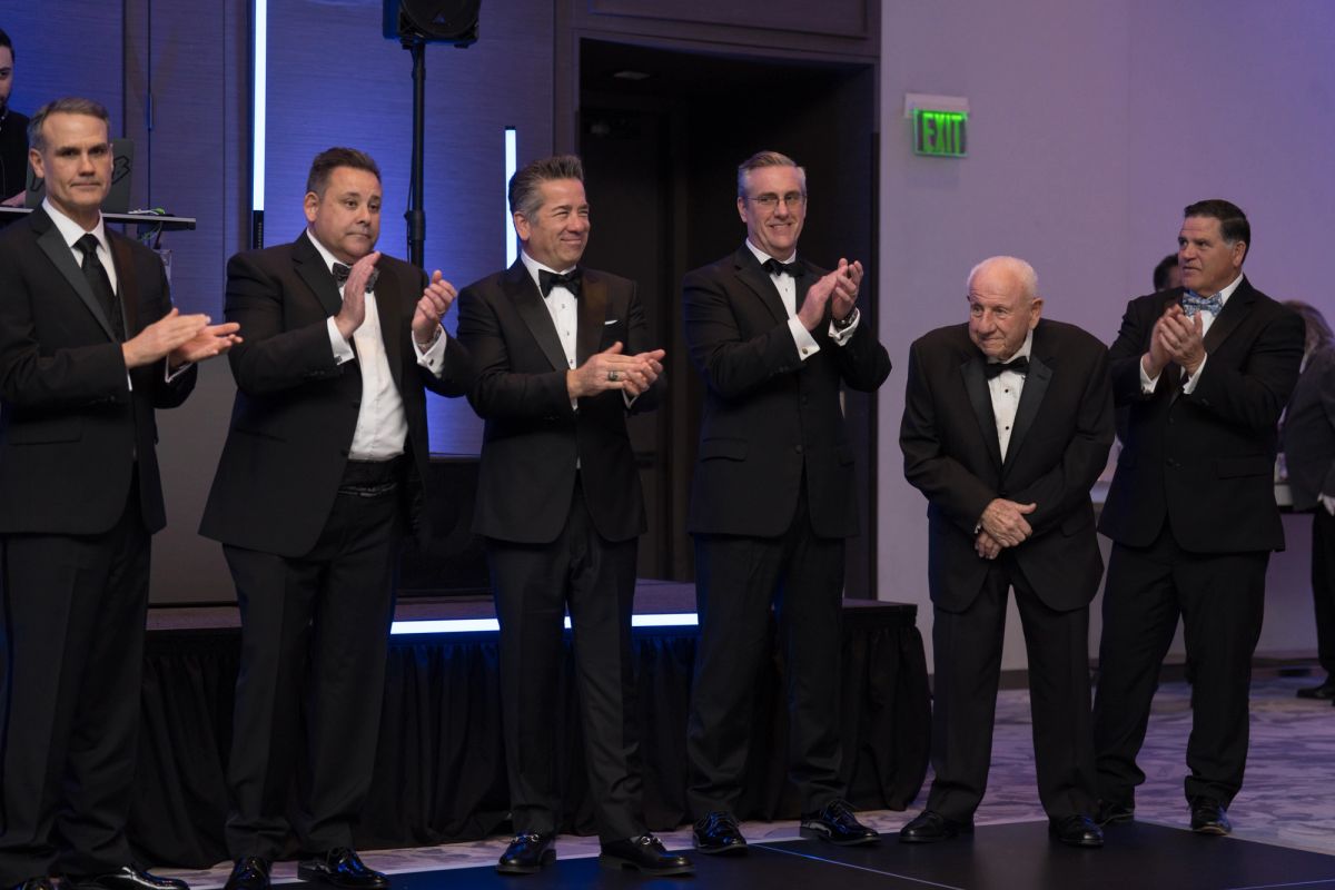Six men in formal tuxedos applauding on stage at an elegant event venue with purple lighting.