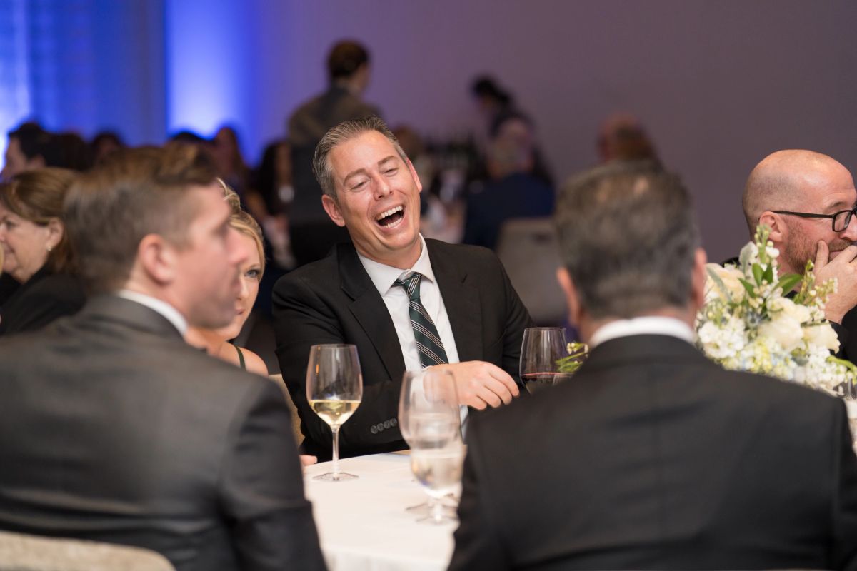 Man in suit laughing heartily at formal dinner table with other attendees in suits at elegant event venue