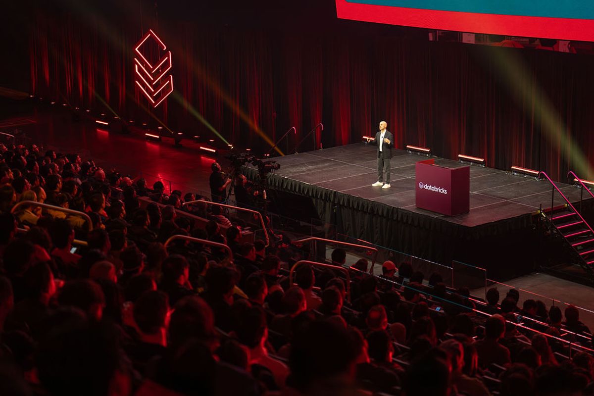 Speaker on stage addressing large audience in red-lit auditorium with geometric logo backdrop