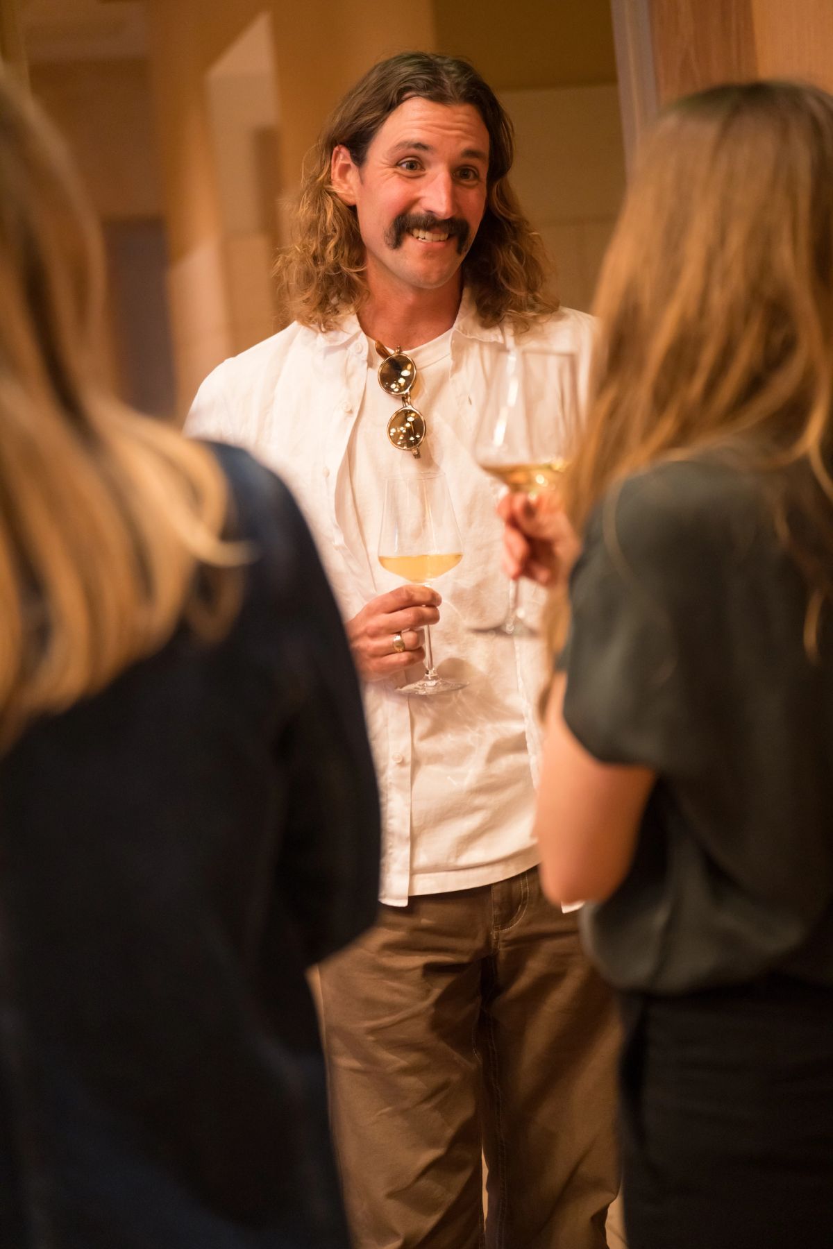 Man with long hair and mustache holding wine glass talks with attendees at networking event