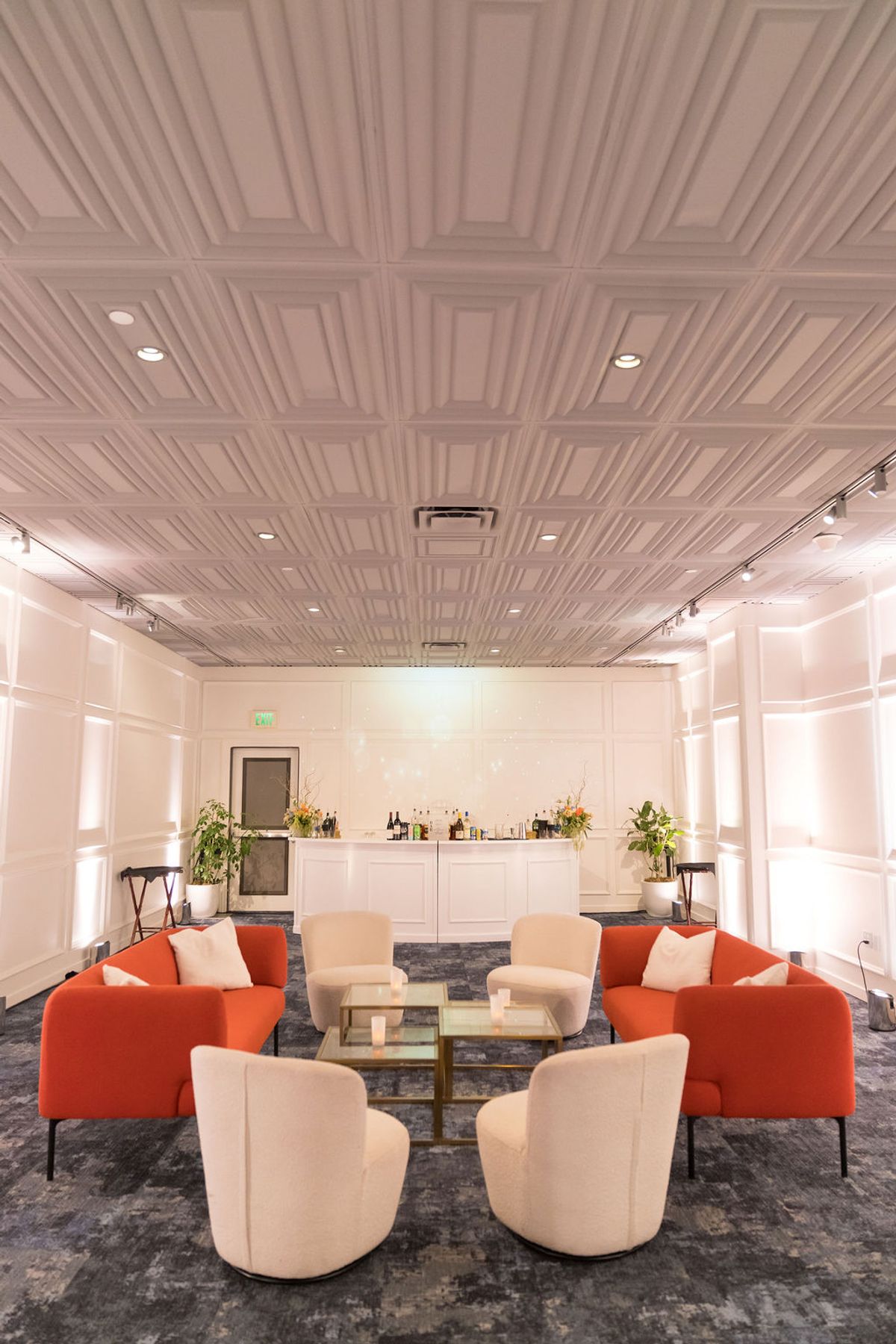 Elegant lounge area with orange and beige seating arranged around glass coffee table, ornate ceiling and.