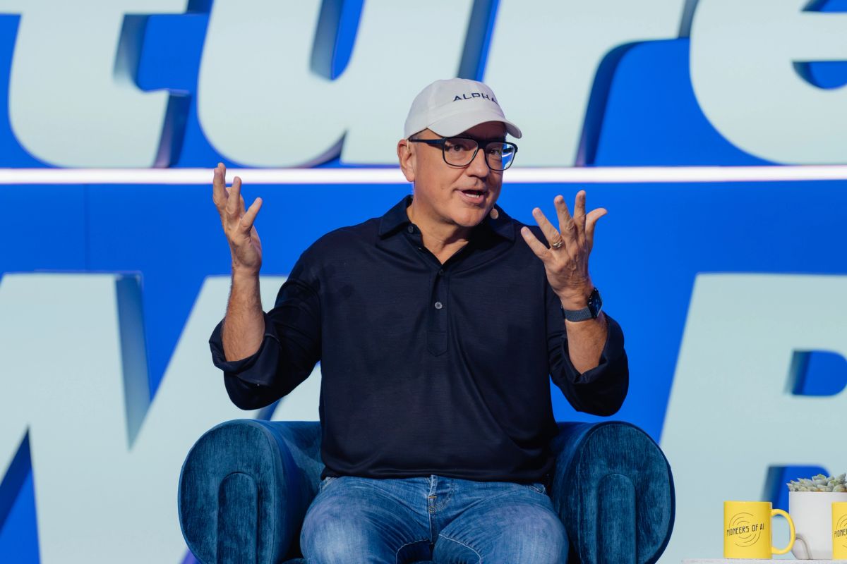 Speaker in black polo and baseball cap gestures with both hands while seated in blue chair on stage