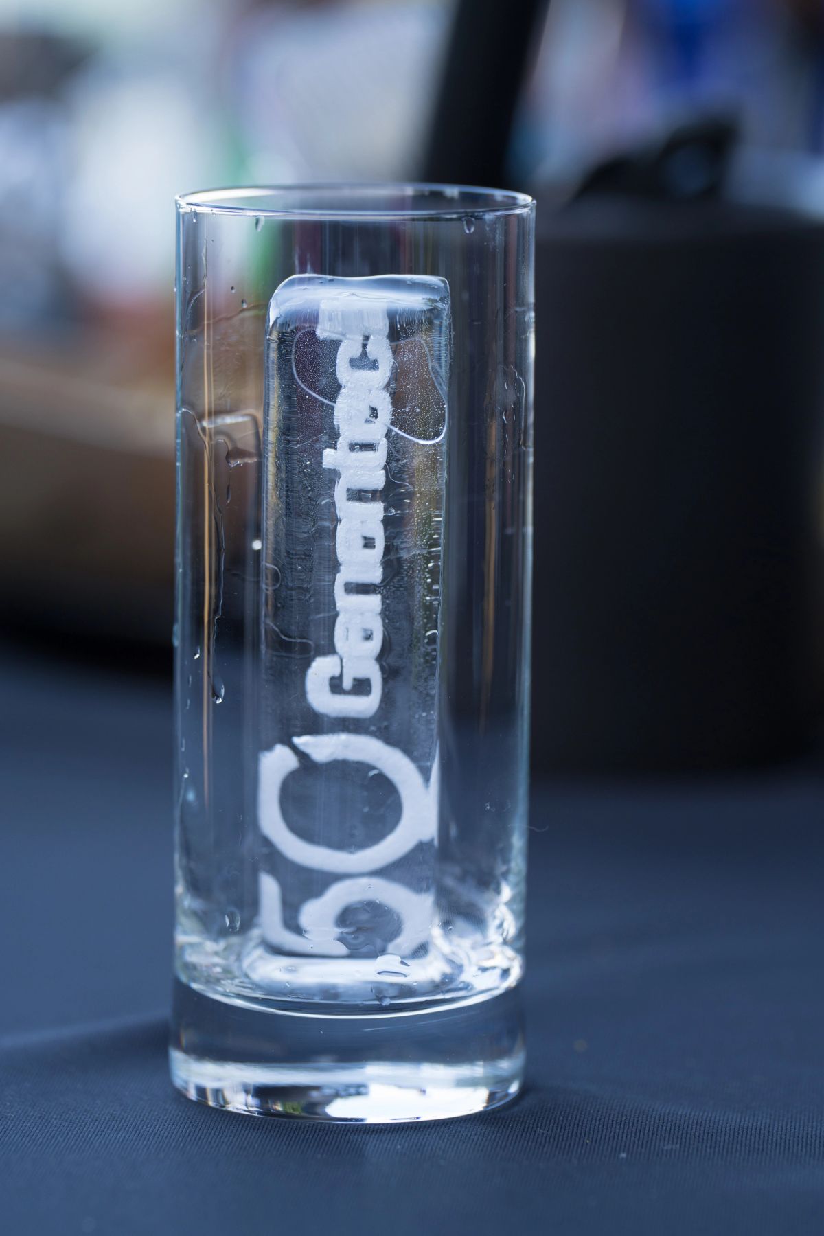 Clear glass with "Genentech" text sits on dark surface with blurred conference room background