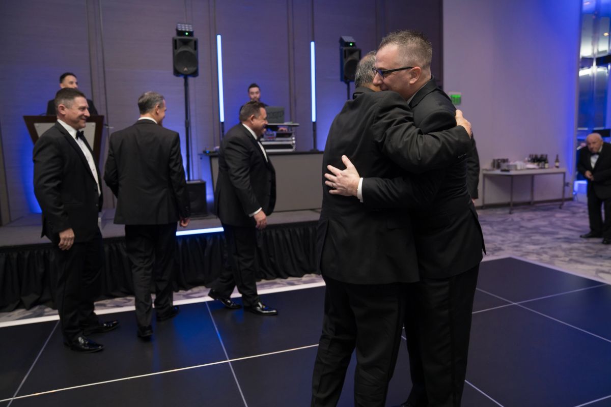 Two men in black suits embracing while other formally dressed attendees mingle in an elegant event space with.