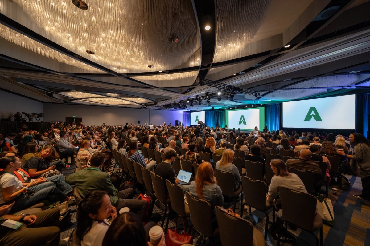 Large conference audience seated in ballroom, facing screens displaying green "A" logo and stage speaker.