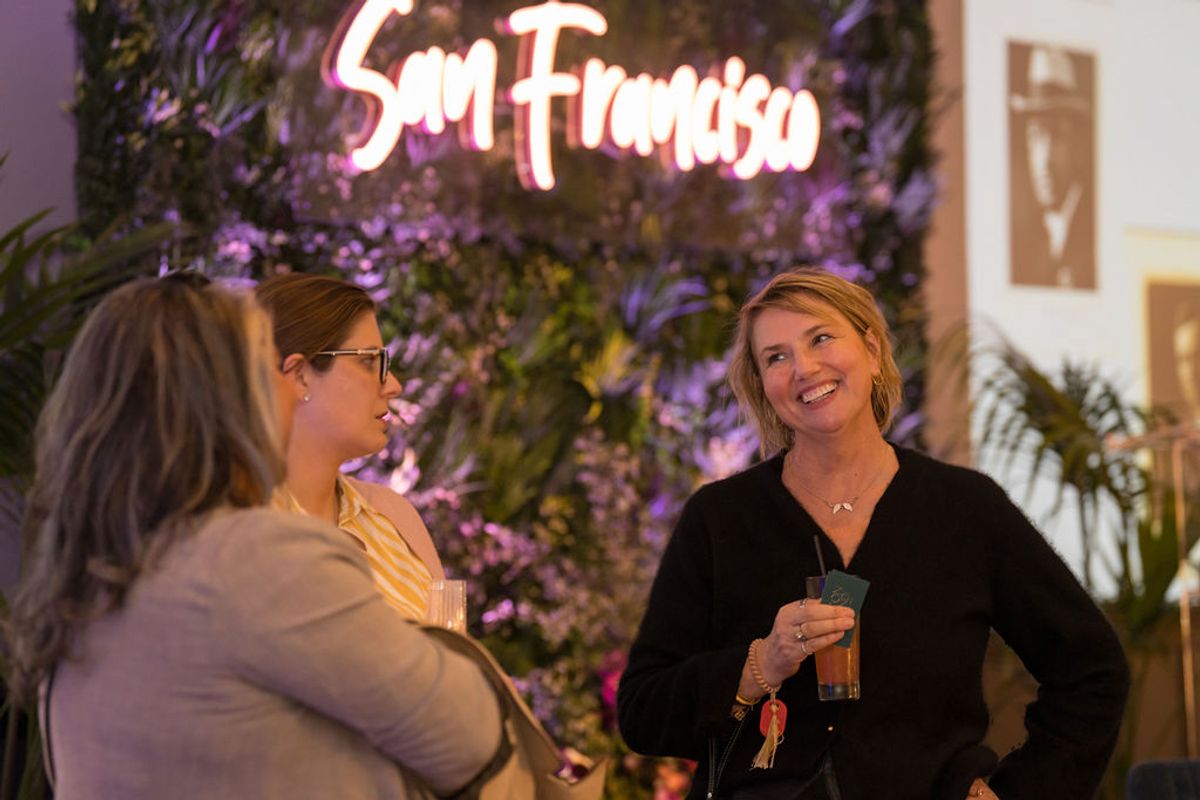 Three women networking and socializing at an indoor reception with neon signage and plants in background.