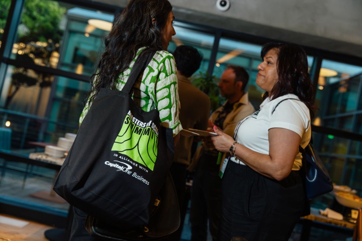 Two women networking at an indoor conference event, one carrying a black tote bag with green design