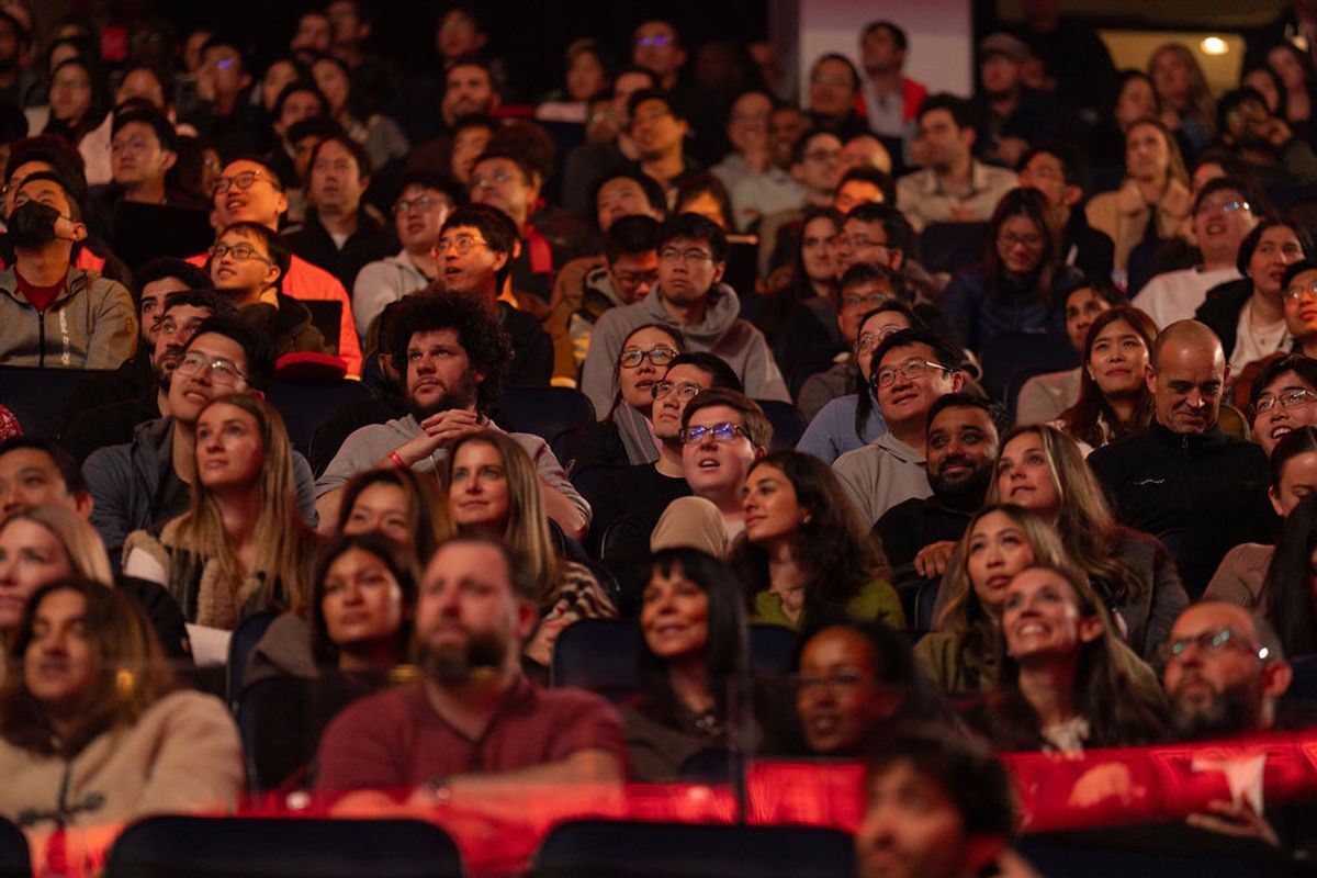 Large audience seated in tiered theater seating, looking upward with engaged expressions during a presentation