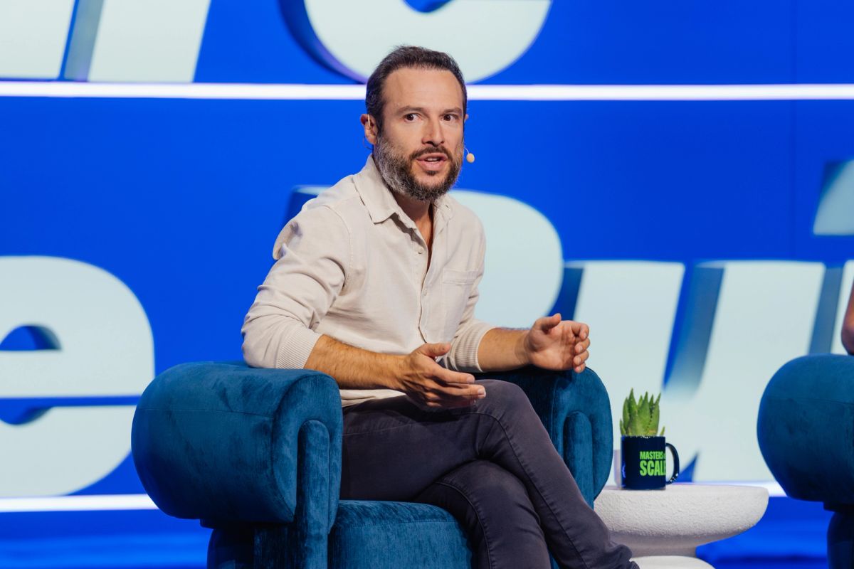 Speaker in beige shirt sitting in blue armchair on stage with blue backdrop at conference event