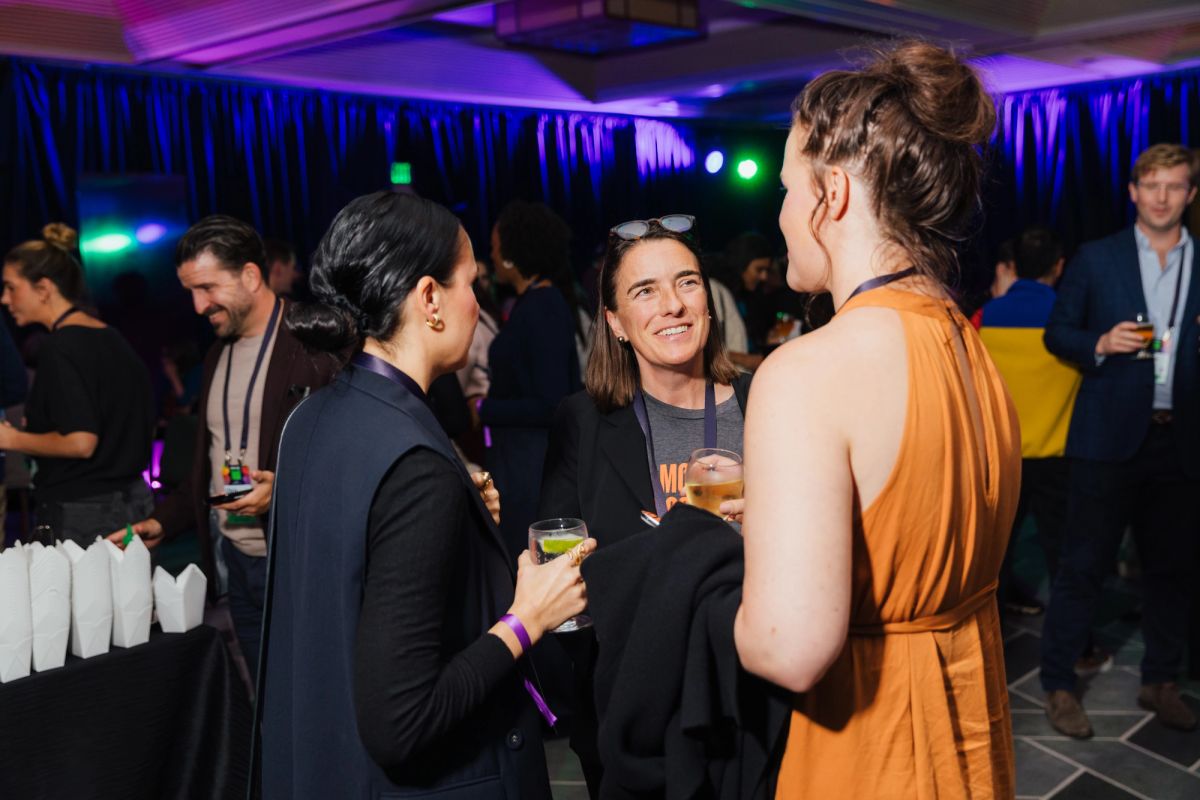 Three women networking with drinks at an indoor conference event with purple lighting and decorations
