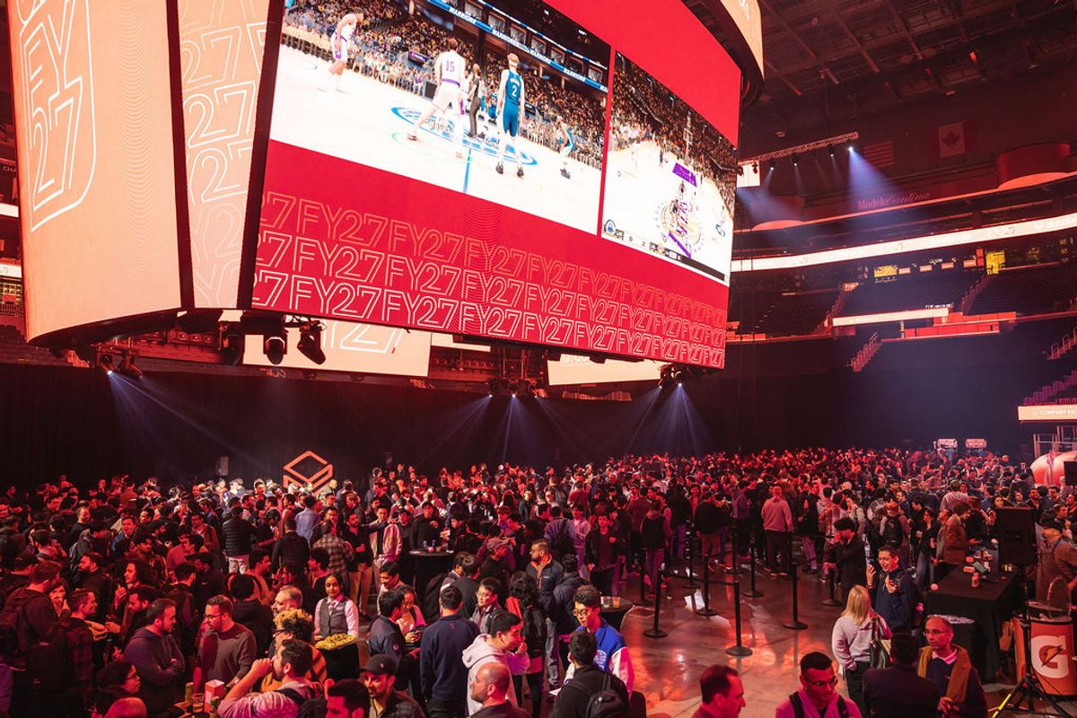 Large crowd of attendees gathered in arena venue with massive overhead screens displaying event content
