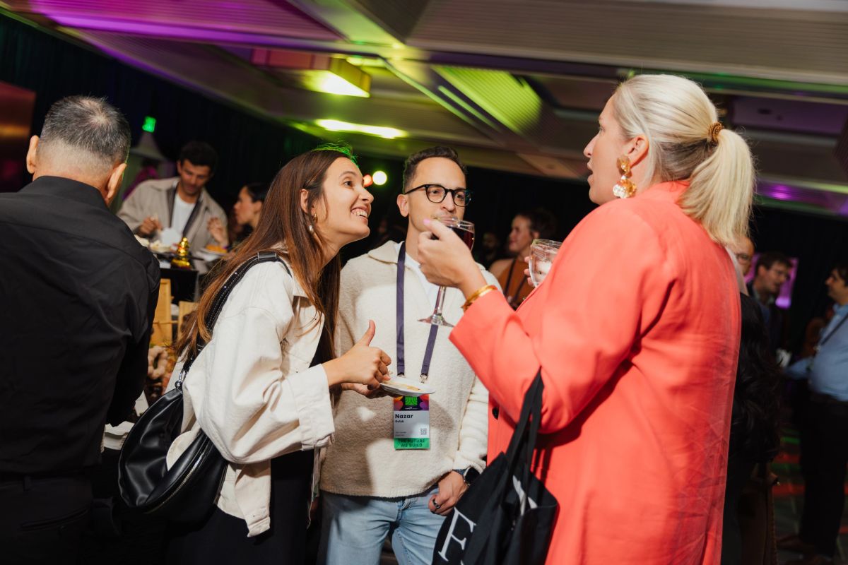Three attendees networking and conversing at an indoor conference event with colorful lighting and crowd in.