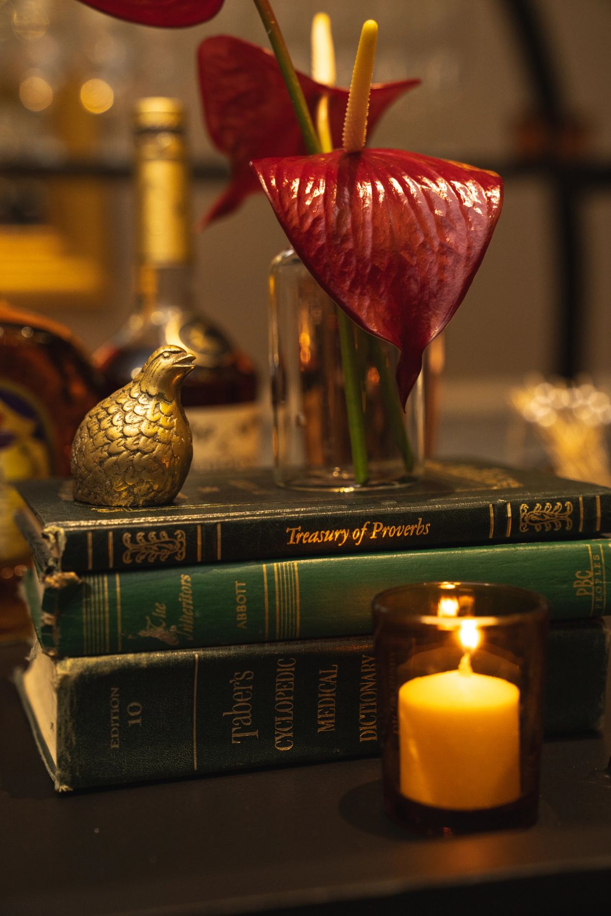Stack of books including "Treasury of Proverbs" with red anthurium flowers, decorative bird, and lit candle