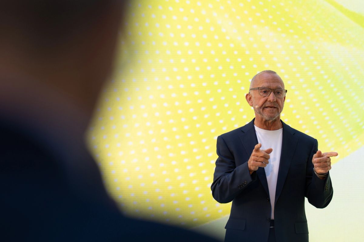 Older male speaker gesturing while presenting in front of yellow dotted backdrop.