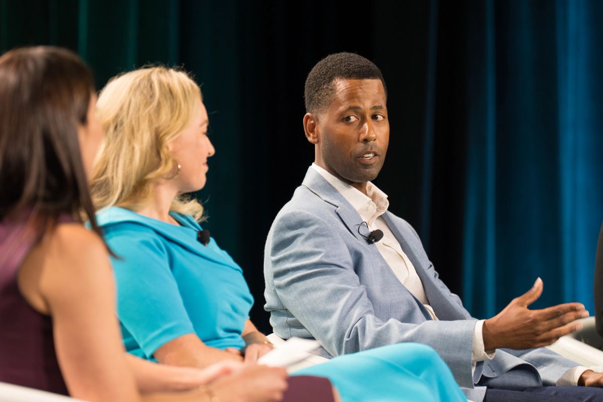 Panel discussion with three seated speakers, one man gesturing while speaking at a conference.