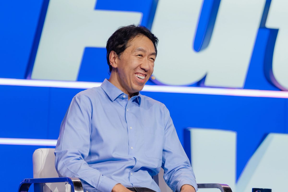 Speaker in blue shirt sitting and smiling on stage with blue backdrop at conference event.