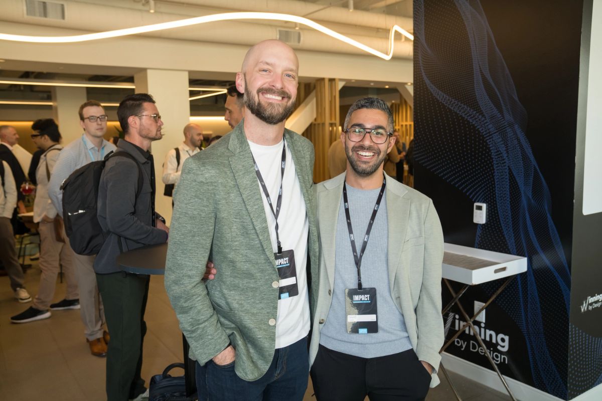 Two conference attendees with name badges smile at a networking event with other professionals in background