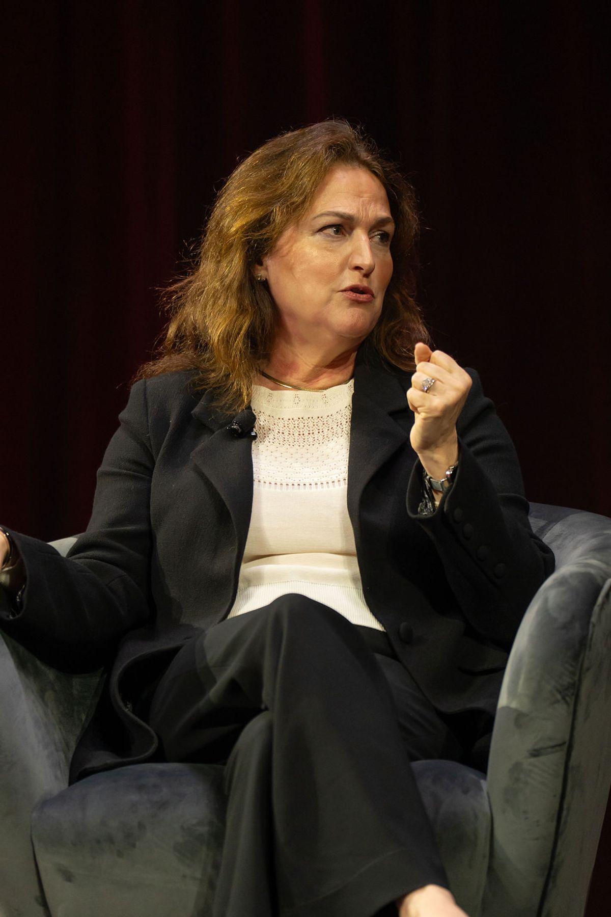Woman in dark business suit speaking at conference, seated in gray chair against dark backdrop