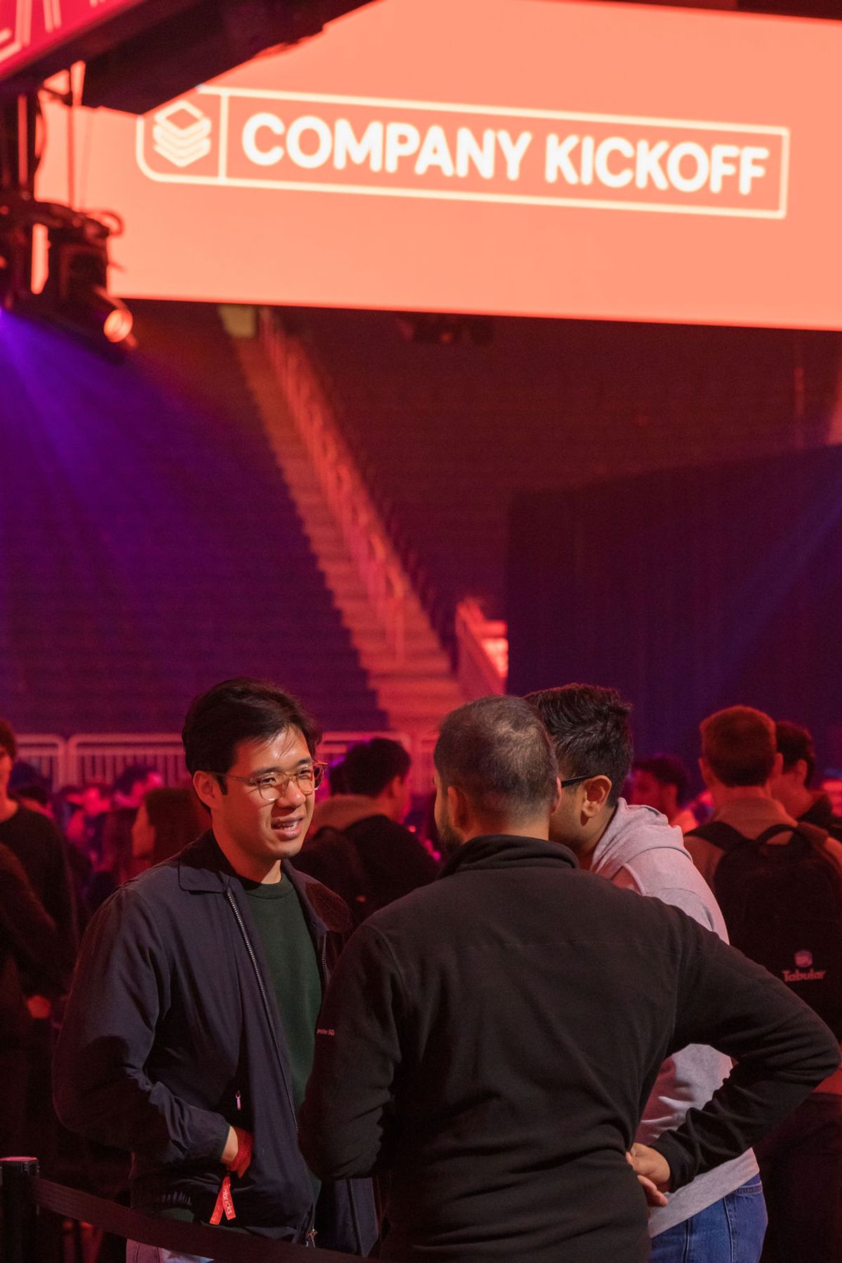 Attendees networking at company kickoff event with red and purple stage lighting in background