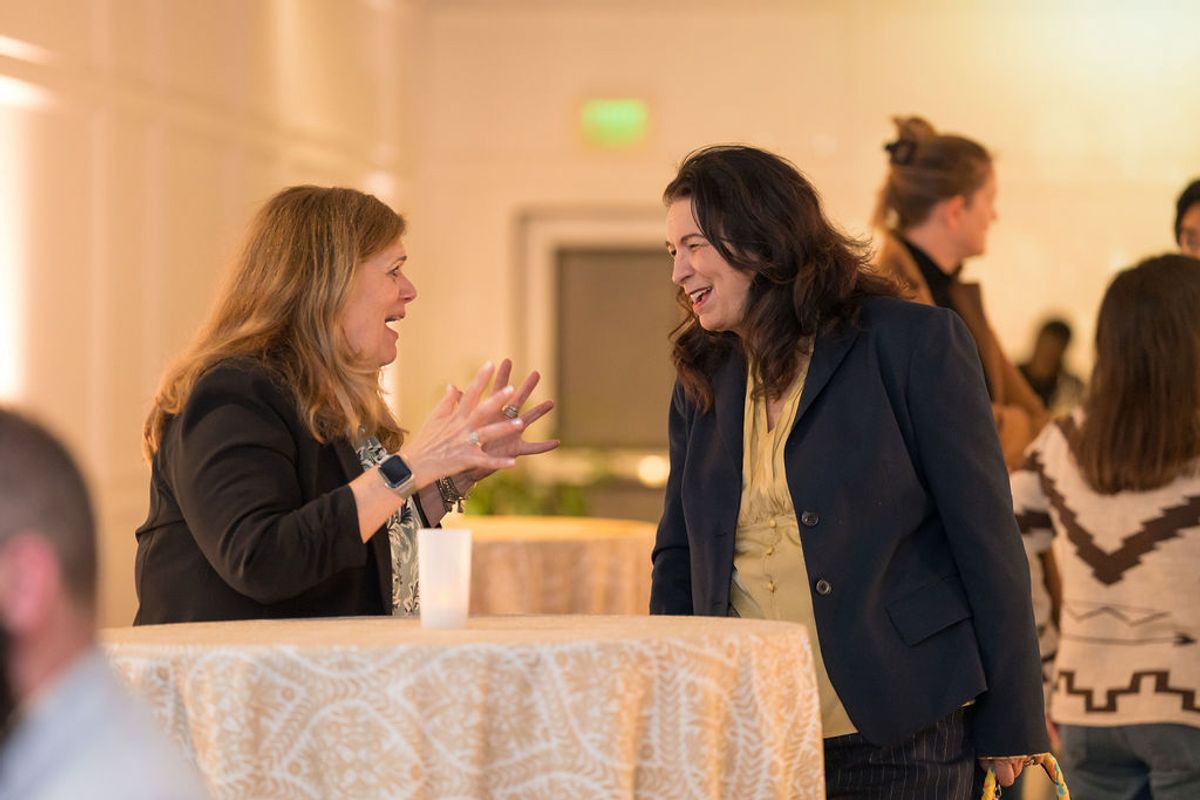 Two women in business attire having an animated conversation at a networking reception with other attendees.