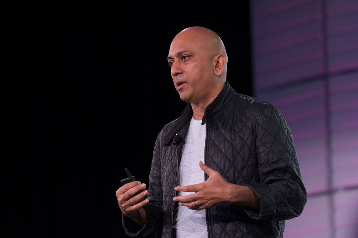Bald speaker in black quilted jacket presenting on stage with purple lighting in background