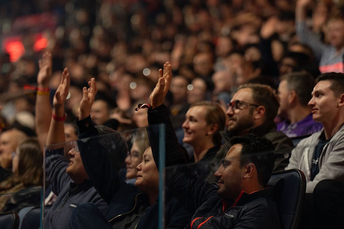Audience members raising hands and applauding during a conference presentation at an arena venue