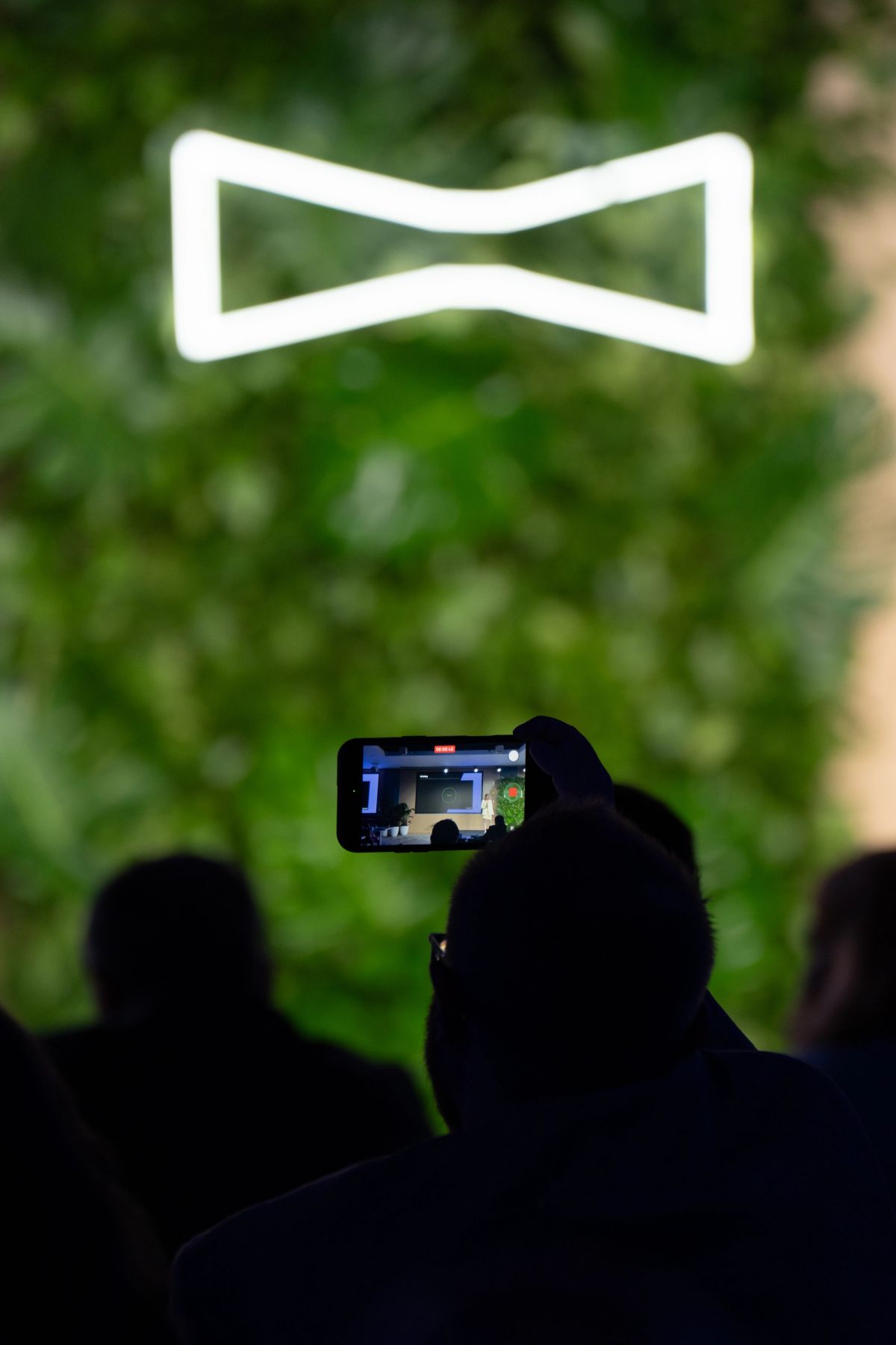 Attendee holds up smartphone to record speaker at conference event with blurred green foliage backdrop