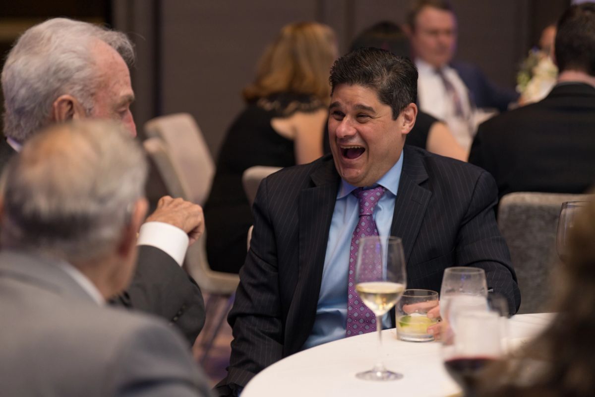 Man in dark suit laughing while seated at dinner table with wine glass, other formally dressed guests visible.