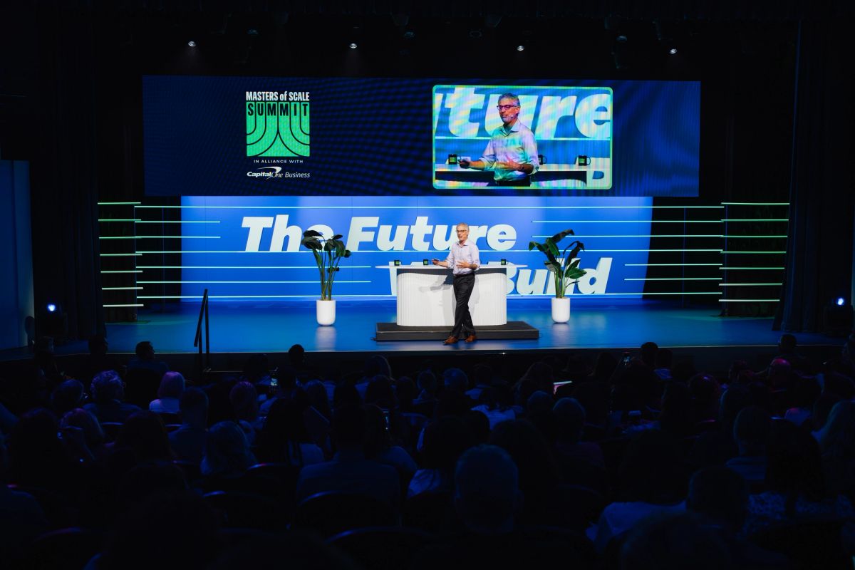 Speaker presenting on stage with "The Future" and "Masters of Scale Summit" displayed on screens to seated.