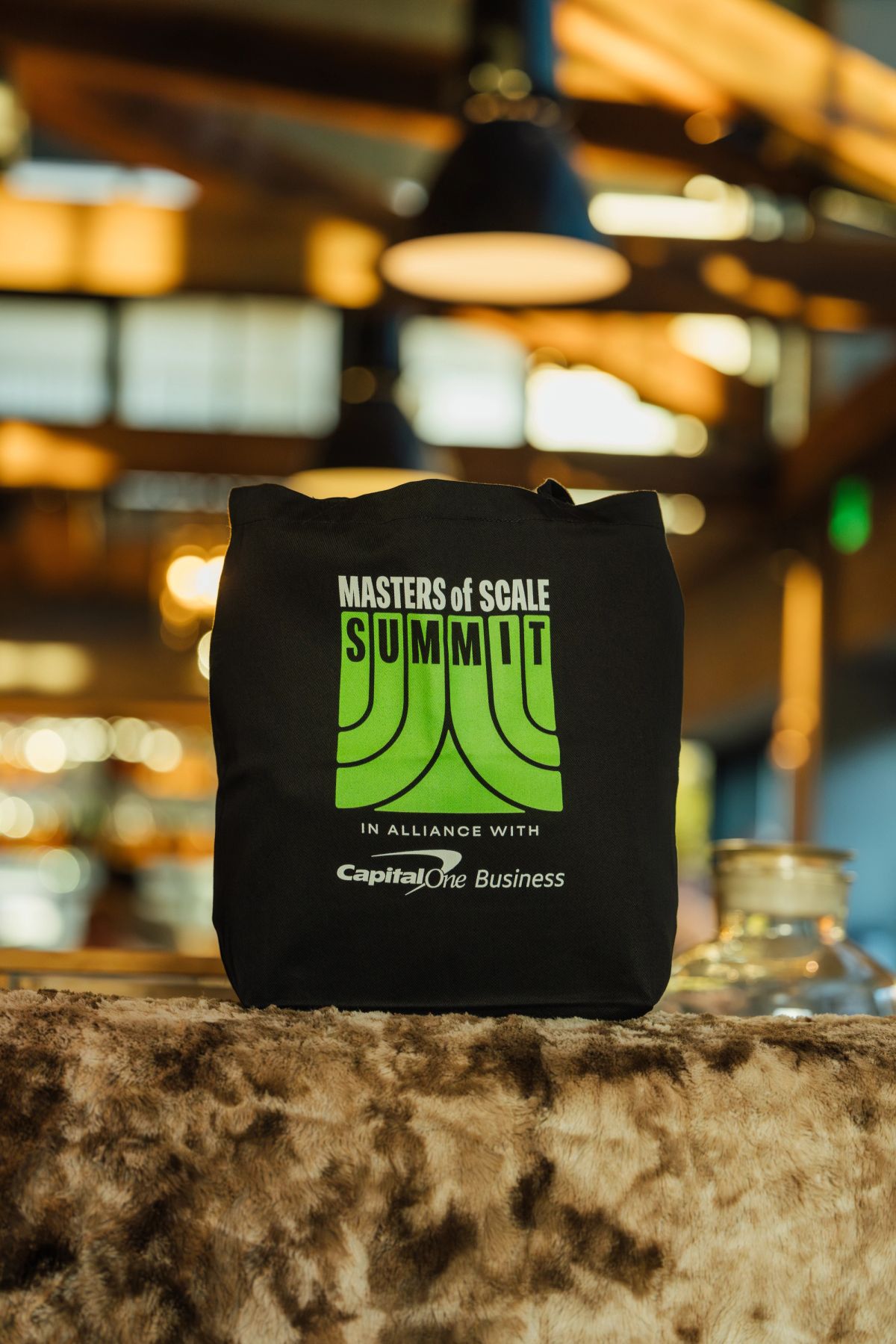 Black tote bag with "Masters of Scale Summit" green logo sits on furry surface in blurred restaurant