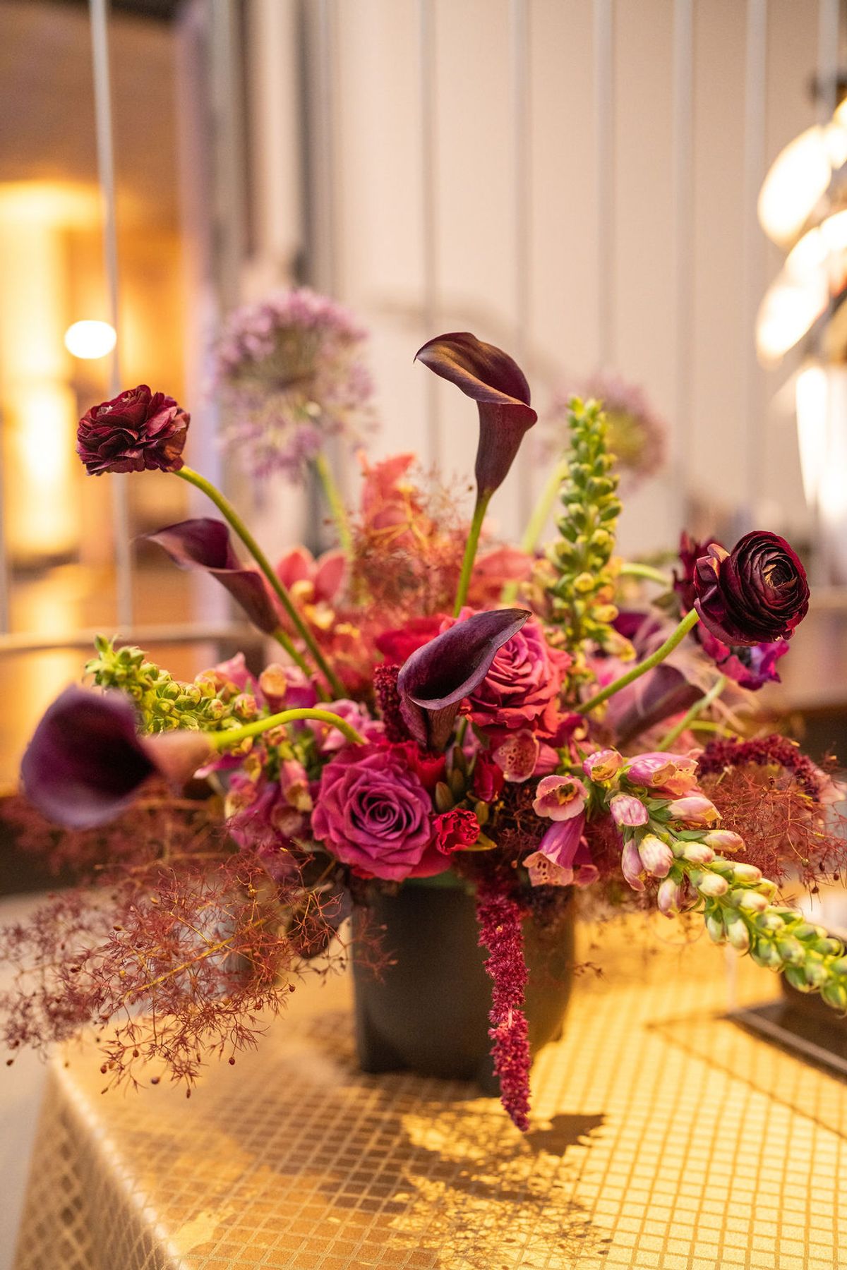Elegant floral centerpiece with deep purple roses, calla lilies, and colorful blooms on golden tiled surface
