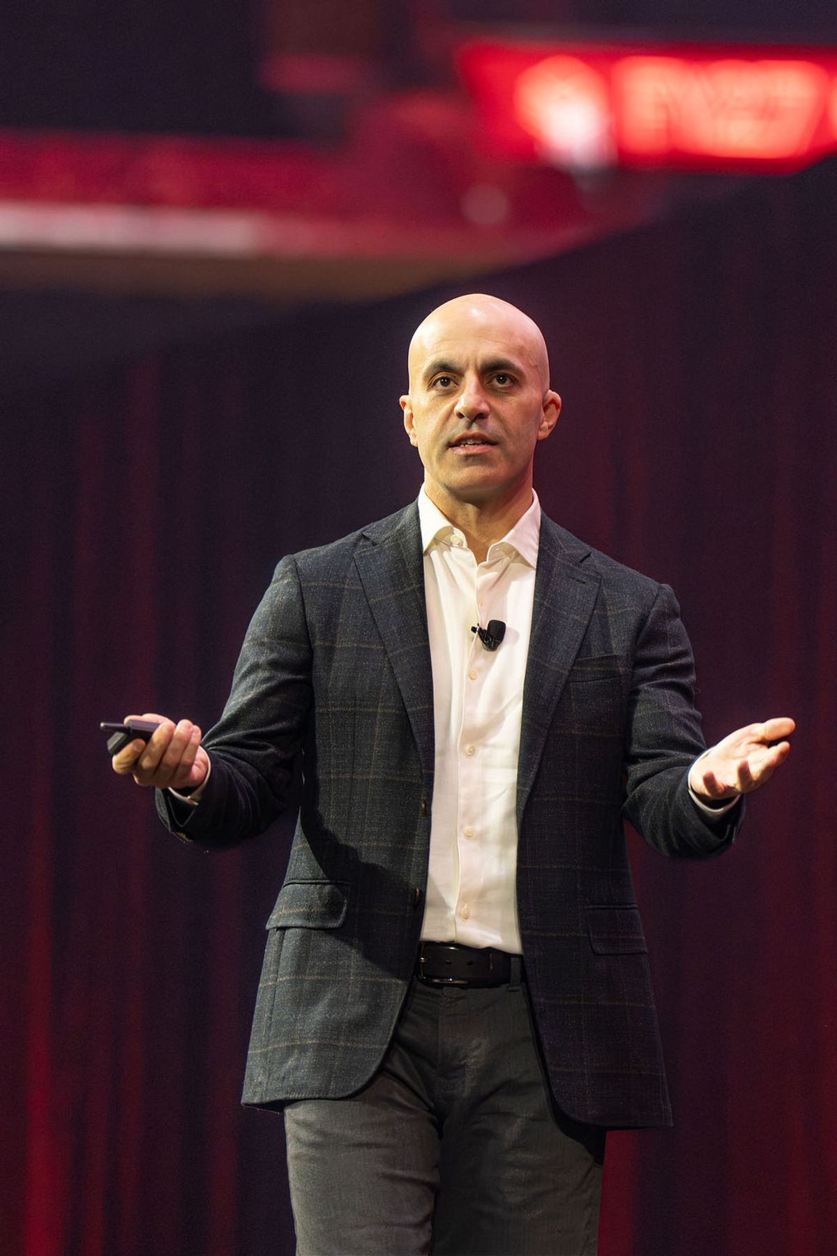 Bald speaker in gray suit gestures with both hands while presenting on stage with red lighting in background