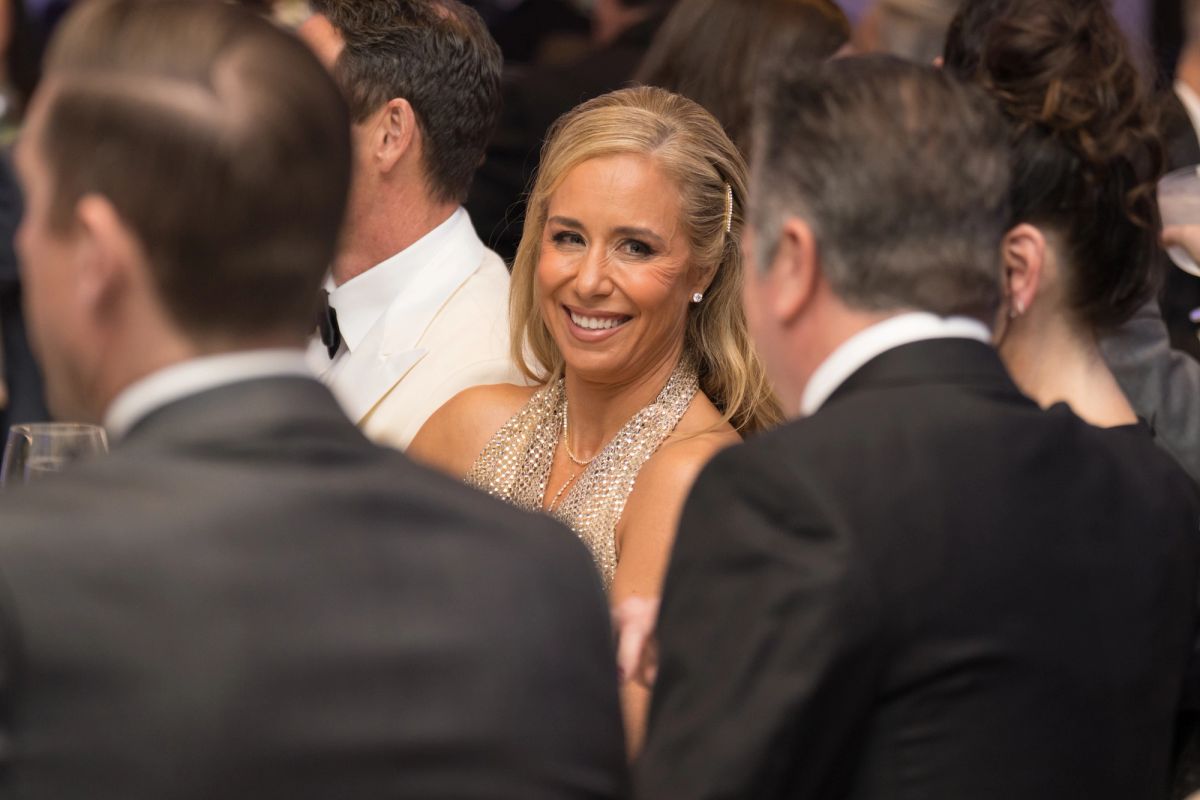 Woman in beaded dress smiling at formal event surrounded by men in suits at crowded reception