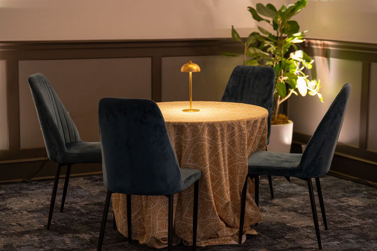 Round dining table with tablecloth, four dark upholstered chairs, brass table lamp, and potted plant in.