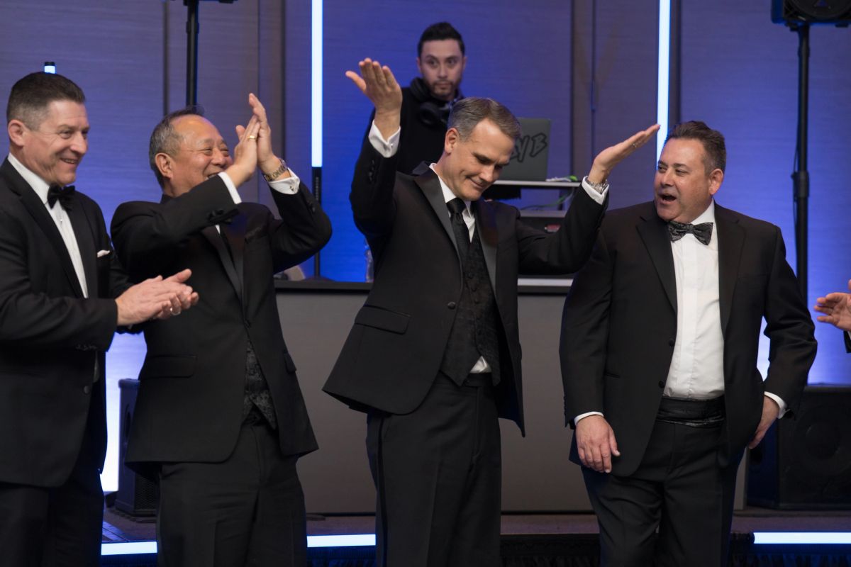 Five men in formal tuxedos celebrating with raised arms and high-fives on a stage with blue lighting