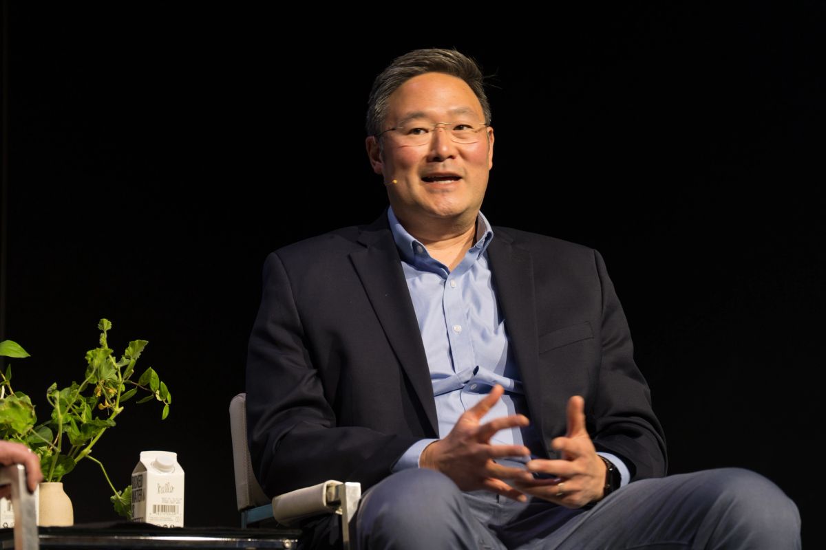 Business executive speaking at conference panel, seated in chair with hands gesturing, plant and water bottle.