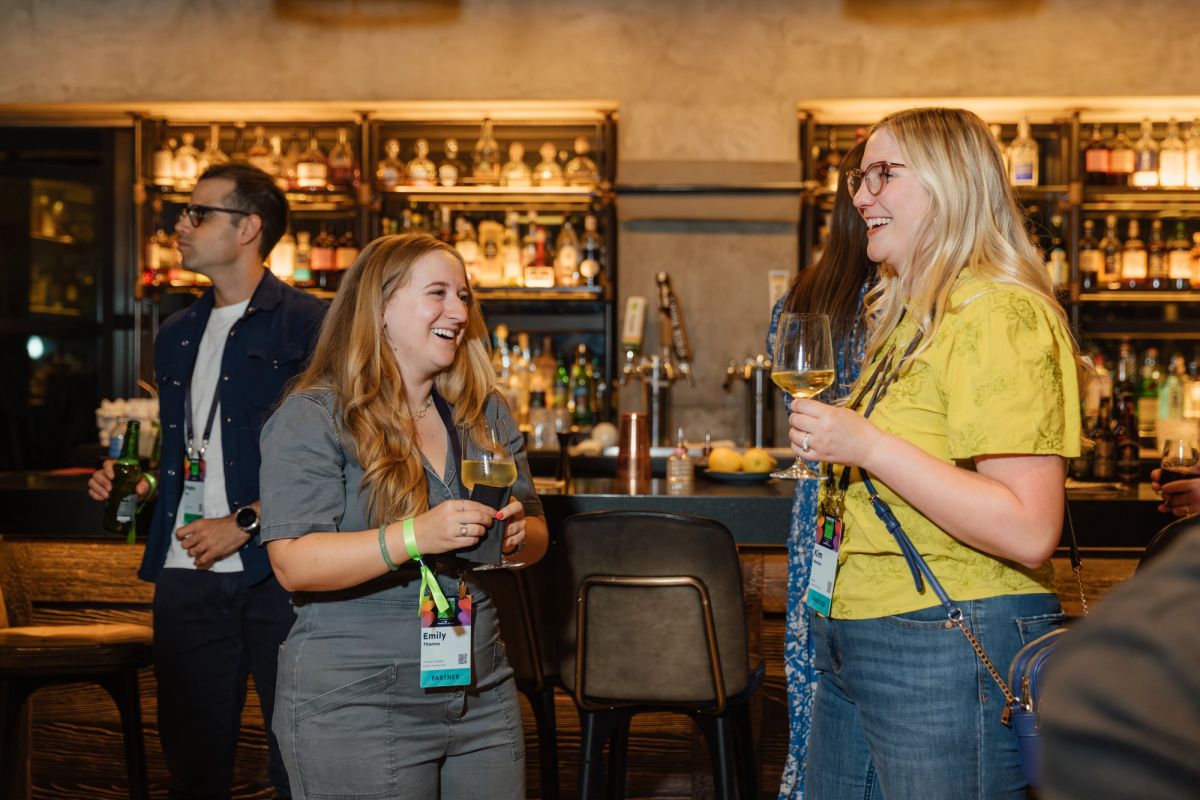 Three conference attendees with name badges socializing at a bar, holding drinks and laughing together