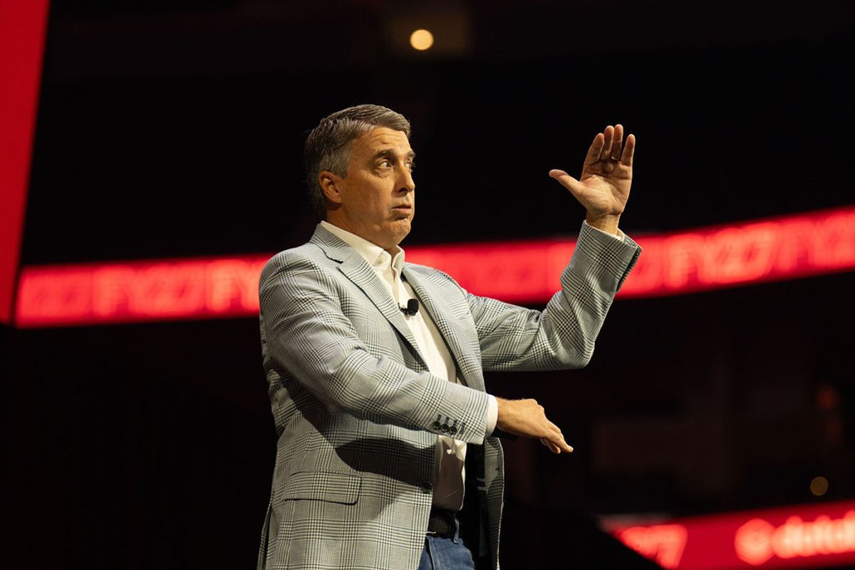 Speaker in gray suit gesturing while presenting on stage with red lighting and backdrop