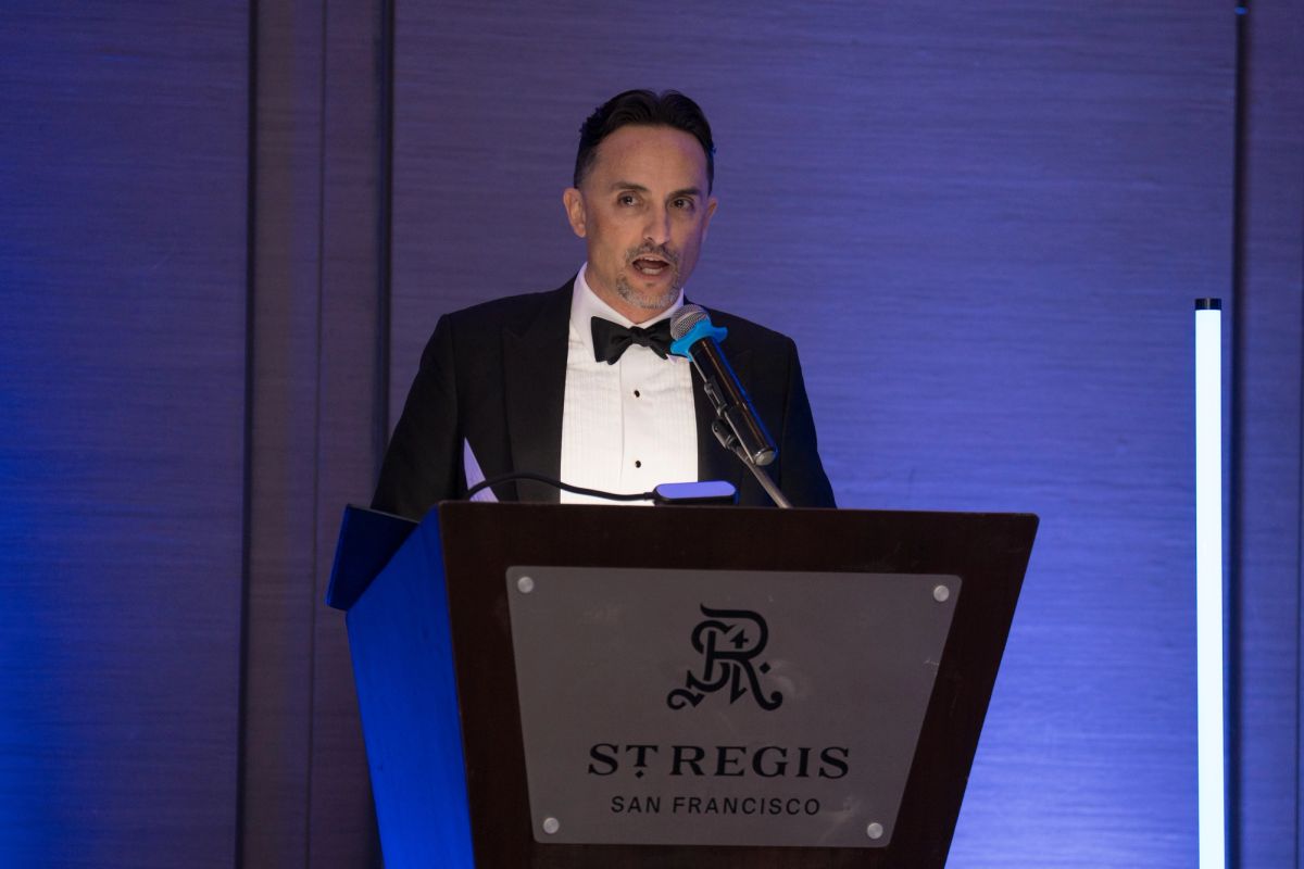 Speaker in black tuxedo at podium with St. Regis San Francisco logo during formal gala event