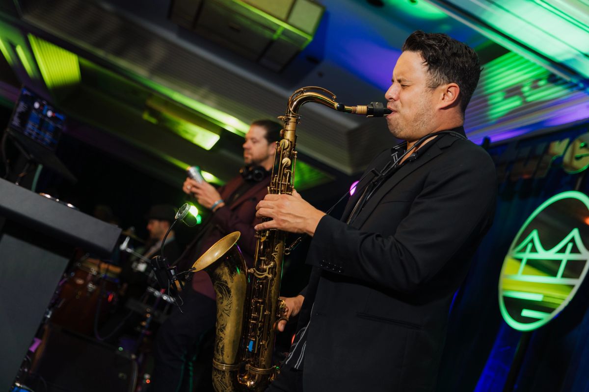 Musician in black suit plays saxophone on stage with colorful lighting and another performer in background