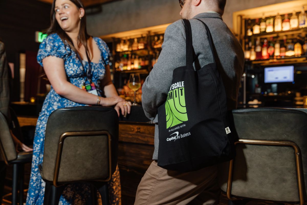 Two people networking at a bar, one wearing a black tote bag, during a conference event