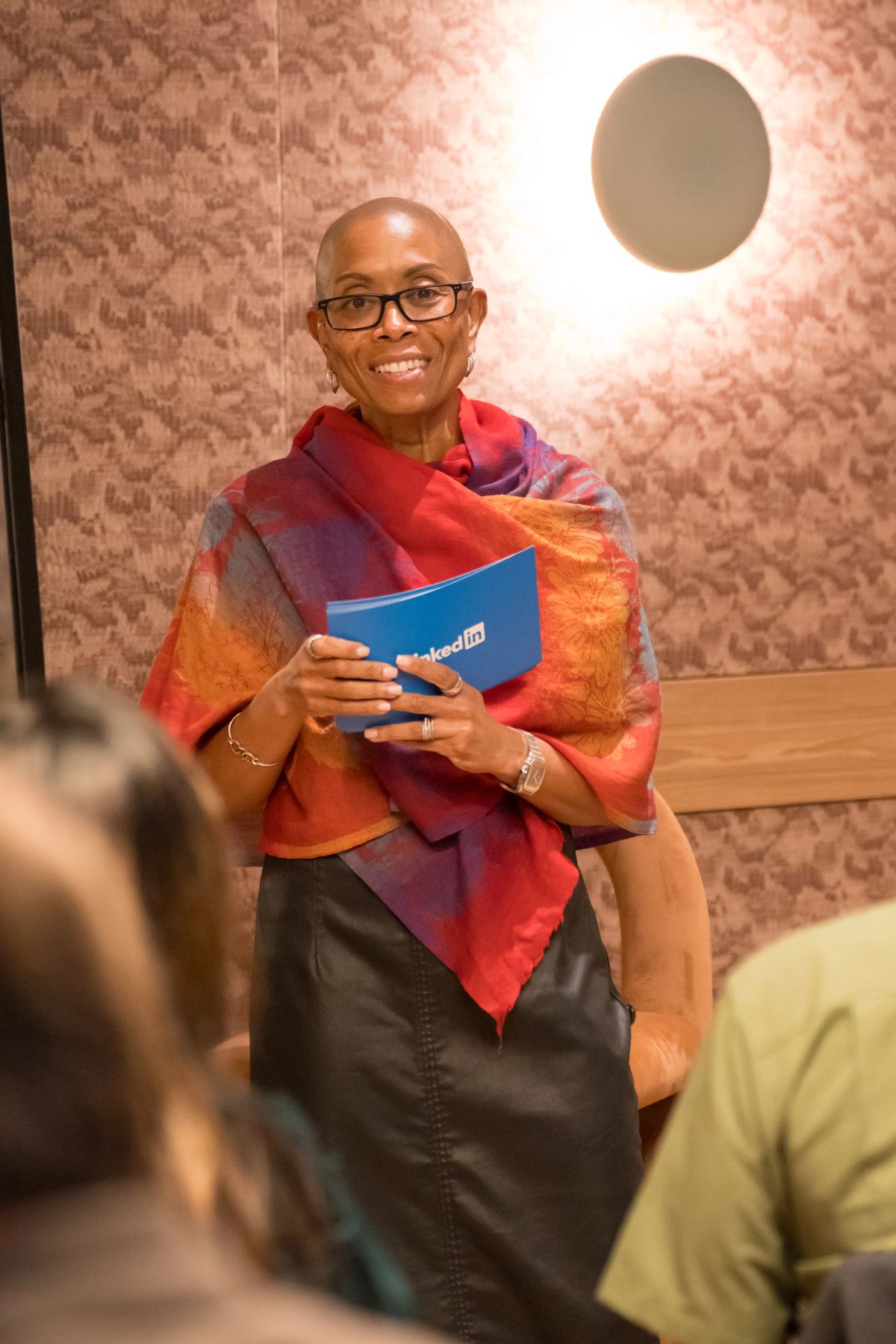 Smiling woman in colorful scarf and glasses holds blue LinkedIn folder while seated at panel discussion