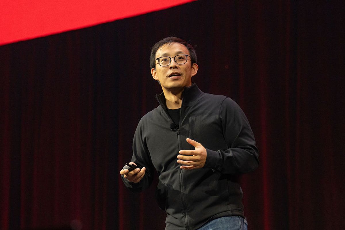 Speaker in dark jacket and glasses presenting on stage with red and black backdrop