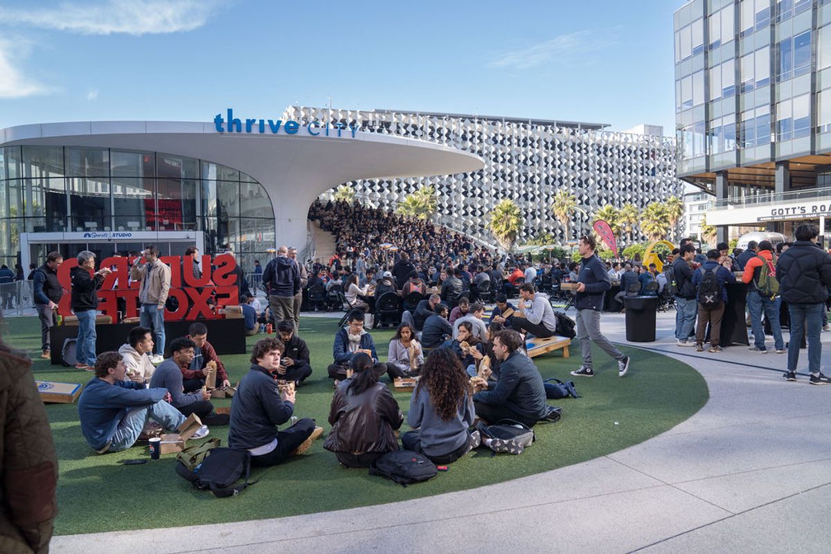Conference attendees sit on artificial turf in outdoor plaza area with modern buildings in background
