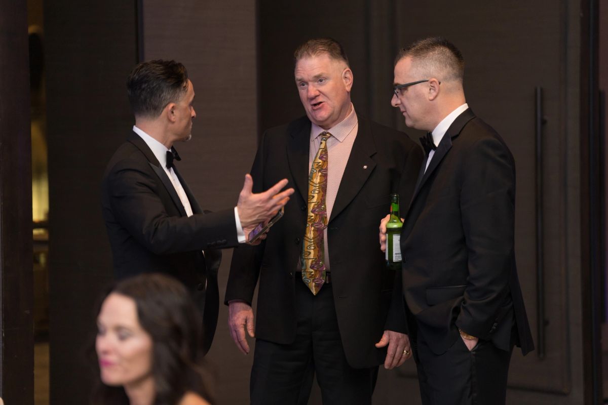 Three men in formal suits having a conversation at an upscale evening event, one holding a drink