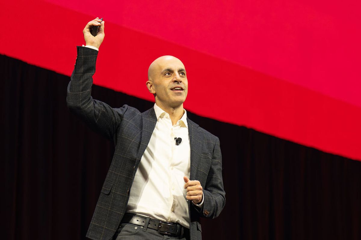 Bald executive in gray suit presenting on stage, holding up small device, with red and black backdrop